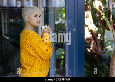 Thoughtful fashionable unaltered biracial woman holding coffee, looking out of window, copy space. Relaxation, free time and domestic lifestyle. Stock Photo