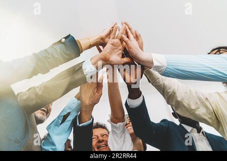 Low-angle shot of a diverse team of colleagues standing together in a bright office, giving a group high five. Focus is on the hands, with light flare Stock Photo