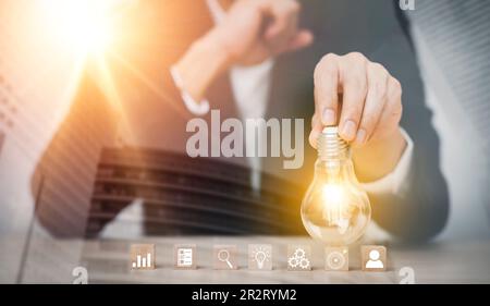 Businessman hand holding light bulb with icon on wood cube and   working on desk, Creativity and innovation are keys to success.Concept of new idea an Stock Photo