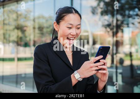 Young business woman surprised and excited watching the success with her stock shares with a cellphone app. Happy startup corporate female executive wearing formal suit smiling using a smartphone. High quality photo Stock Photo