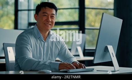 Mature Asian businessman chinese korean office worker specialist male work on computer develop project online look at camera confident smile. Portrait Stock Photo