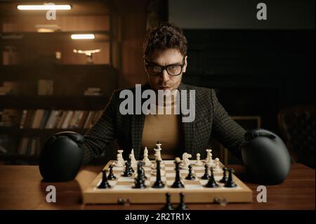 Angry young man wearing boxer gloves sitting at table with chessboard Stock Photo