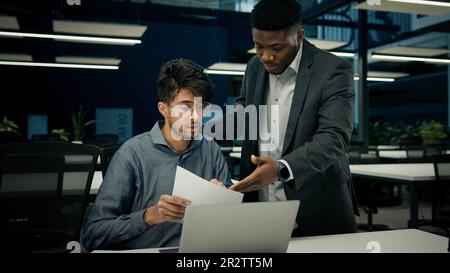 Male African boss businessman mentor teaching indian man trainee new employee discuss online business project on laptop explain details documents to Stock Photo