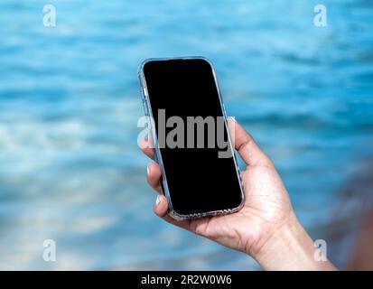 Mobile beach phone with empty screen. Mockup image of black blank desktop screen in hand holding mobile smart phone on the blue sea background in summ Stock Photo