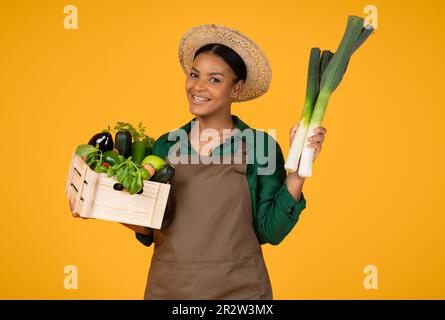 Black Lady Holding Leek And Box With Vegetables, Yellow Background Stock Photo