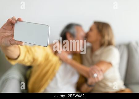 Loving aged caucasian wife kissing husband, man making selfie on smartphone, sitting on sofa, selective focus Stock Photo