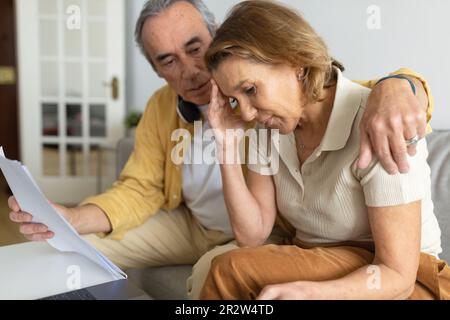 Stressed senior spouses checking financial papers at home, suffering financial crisis, sitting on couch Stock Photo
