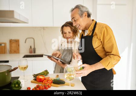 Cheerful senior couple cooking and using digital tablet, looking for nice recipe on Internet in kitchen at home Stock Photo