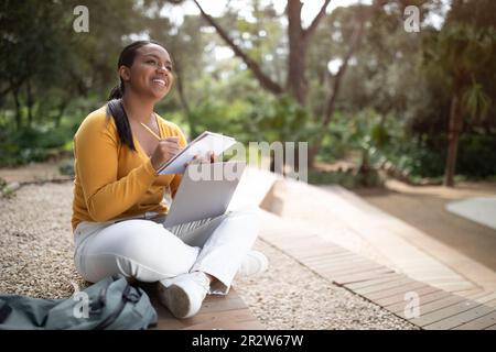 Cheerful brazilian lady attending webinar outdoors, sitting in park, using laptop and taking notes, copy space Stock Photo