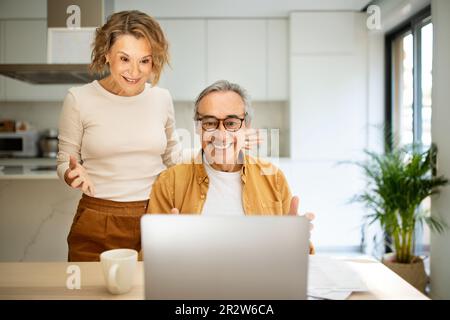 Happy senior spouses man and woman sitting at kitchen table in front of laptop computer, looking at screen and smiling Stock Photo