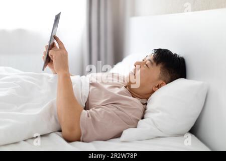 Happy asian man using digital tablet in bed, side view Stock Photo