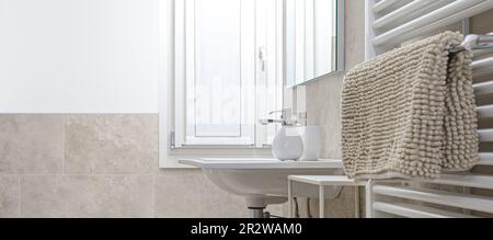 Modern bathroom. Sink with soap and towel on white radiator. Toilet furnished with marble and beige ceramic tiles. Bright room illuminated by light en Stock Photo