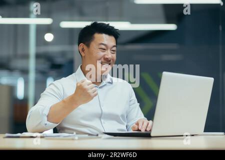 Young Asian businessman working in office at laptop and happy with success, showing victory hand gesture, satisfied with result, successful deal. Stock Photo