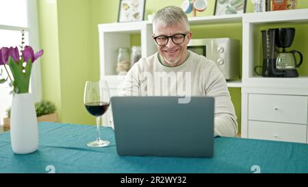 Middle age grey-haired man drinking glass of wine using laptop at home Stock Photo