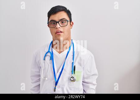 Young hispanic man with down syndrome wearing doctor uniform and stethoscope with serious expression on face. simple and natural looking at the camera Stock Photo