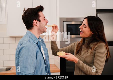 Young couple eating together in affeccionate attitude. Two people laugh and feed each other at home kitchen. Smiling wife flirting giving healthy food to her handsome husband having a romantic lunch. High quality photo Stock Photo