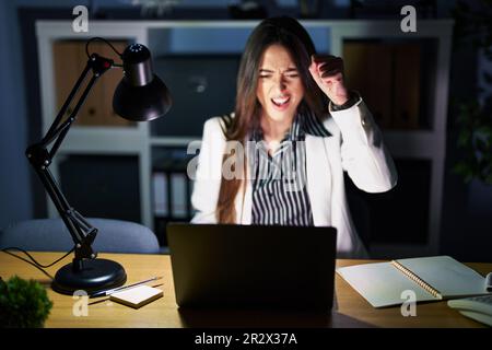 Young brunette woman working at the office at night with laptop angry and mad raising fist frustrated and furious while shouting with anger. rage and Stock Photo