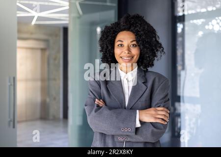 Portrait of successful female boss in business suit, mature African American woman smiling and looking at camera with crossed arms, businesswoman happy with achievement at workplace inside office. Stock Photo