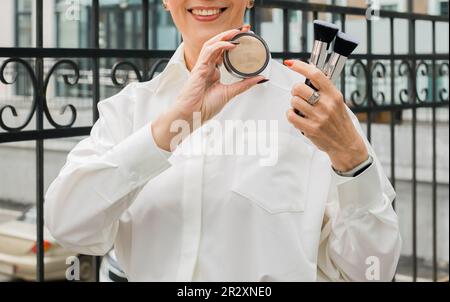 Close up middle aged woman holding jar with loose powder and makeup brushes near face outdoors - make-up artist and cosmetic product for make up Stock Photo