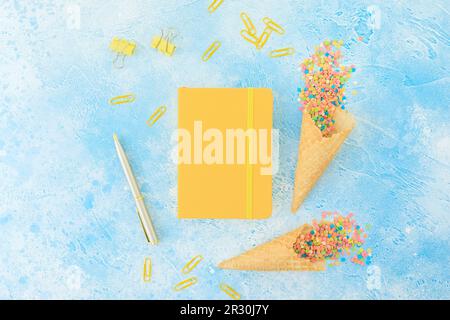 Diary with pen and colorful confetti candy and waffle cone on blue. Flat lay, top view Stock Photo
