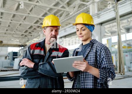Engineers discussing online project on tablet pc while standing in workshop of factory Stock Photo