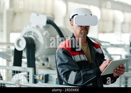 Mature engineer in uniform using digital tablet to watch production of material with VR glasses Stock Photo