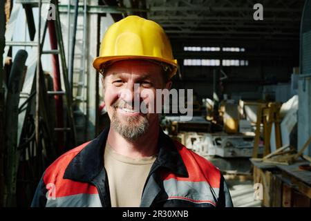 Portrait of mature engineer in work helmet smiling at camera standing in workshop of factory Stock Photo