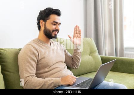 Video call concept. Indian freelancer man waving hello into laptop webcam, guy connected via video with family, friends or colleagues working remotely from home Stock Photo