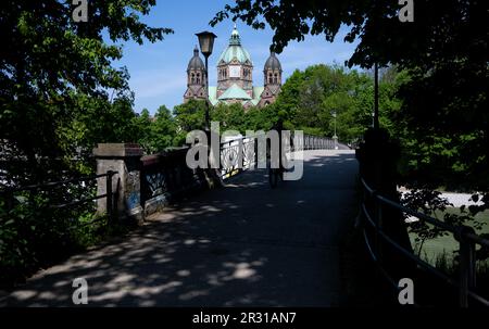 Munich, Germany. 22nd May, 2023. A man rides his bicycle over the cable bridge on the Isar River. The church of St. Luke can be seen in the background. Credit: Sven Hoppe//dpa/Alamy Live News Stock Photo