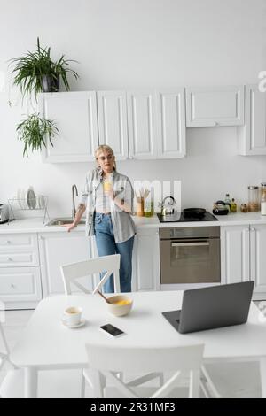 tattooed woman in eyeglasses holding glass of orange juice and standing near desk with devices, bowl with cornflakes and cup of coffee with saucer on Stock Photo