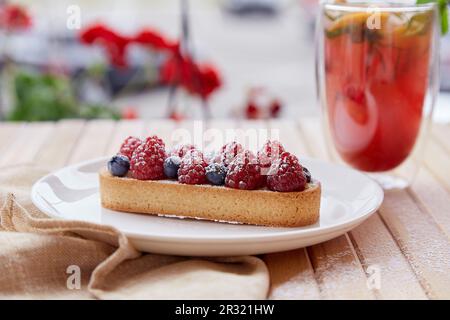 French sugar free tart with raspberry and blueberry at the terrace. Refreshing raspberry - orange lemonade with mint and cane straw outside on the wooden table Stock Photo