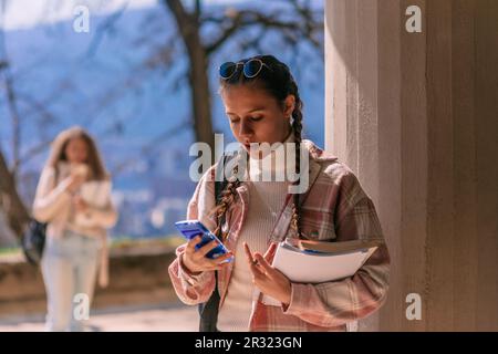 School girl using calculator while holding books and leaning on a pillar at the school yard. Her female friend is chatting on a phone in the back Stock Photo