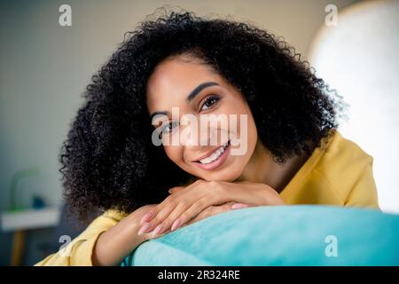 Photo of pleasant toothy beaming candid sincere woman with perming coiffure wear yellow long sleeve smiling on sofa at home room indoors Stock Photo