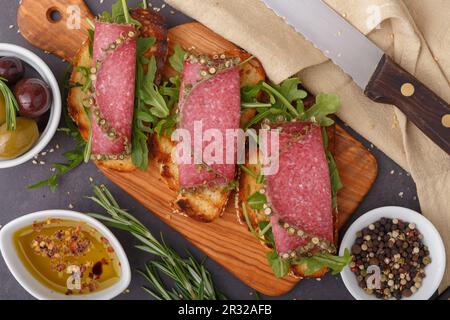 Wholegrain bread with rocket salad and spicy Italian salami on an old grunge rustic wooden board. Dark gray background. Top view. Close up. Copy space Stock Photo
