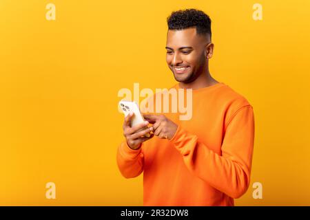 Ecstatic young Indian man standing isolated on yellow and using trendy smartphone, looking at screen and smiling joyfully, millennial guy enjoy new mobile application features Stock Photo