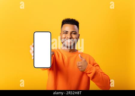 Portrait of handsome young man demonstrates smartphone with blank screen and shows thumb up, guy likes mobile app, looking and smiling at the camera, feels satisfied, isolated Stock Photo