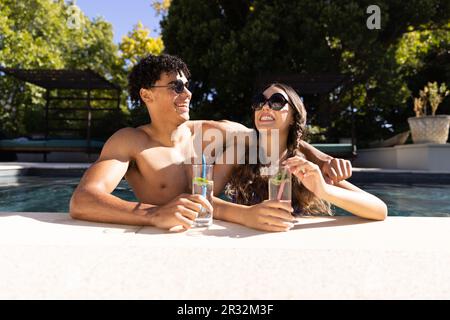 Happy diverse fit couple standing in swimming pool laughing in the sun with cocktails Stock Photo