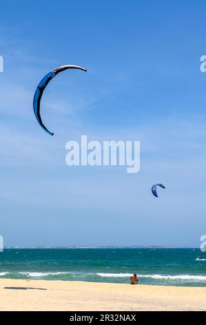 A local man sits alone on the beach observing the kitesurfers in action at sea in Mui Ne, Vietnam. Stock Photo