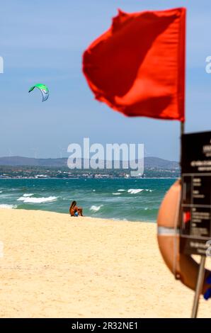 A local man sits alone on the beach observing a kitesurfer in action at sea and 'no swimming' red flag in the foreground in Mui Ne, Vietnam. Stock Photo