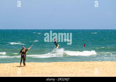 Instructor and learner holding kite lines while a kitesurfer jumps in the air with a twin tip board off the beach in Mui Ne, Vietnam. Stock Photo