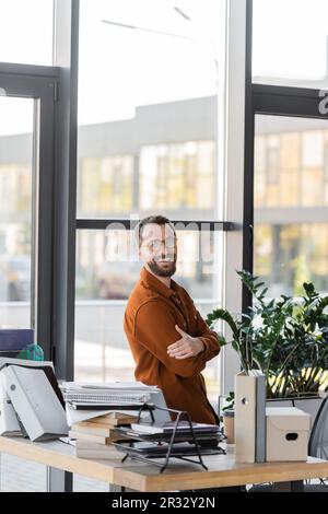 happy bearded businessman in eyeglasses and shirt smiling at camera while standing with crossed arms next to windows and workplace with pile of books, Stock Photo