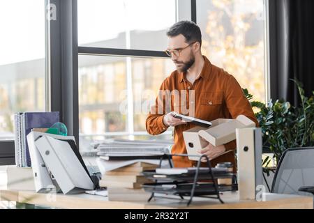 serious bearded businessman in eyeglasses and shirt standing with carton box and notepad next to work desk with folders, pile of books, notebooks and Stock Photo