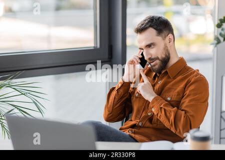 positive and charismatic bearded businessman in stylish shirt talking on mobile phone while sitting near blurred laptop and windows in contemporary of Stock Photo