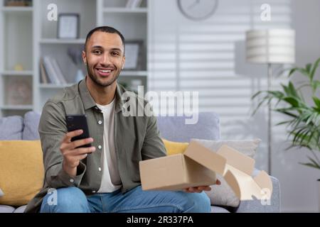 Portrait of a happy young African-American man sitting at home on the sofa ,with a phone in his hands and holding a package, he ordered in an online store with delivery. Stock Photo