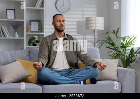Yoga classes at home. Young African American man sitting relaxed on couch in lotus position with eyes closed, meditating, resting. Stock Photo