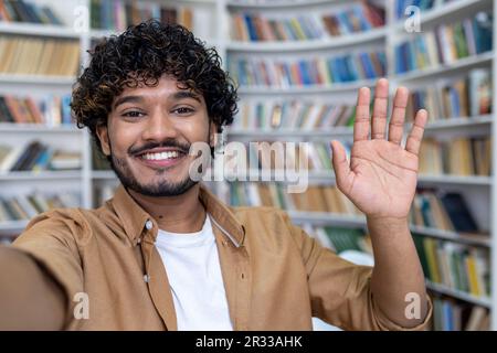 Young hispanic male student chatting online via video call in the library. Smiles at the camera, shoots a blog, waves, studies. Close-up photo. Stock Photo
