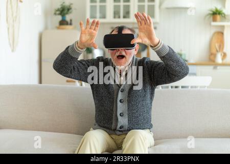 Excited mature senior man wearing using virtual reality metaverse VR glasses headset at home. Grandfather touching air during VR experience on virtual reality helmet. Simulation hi-tech videogame Stock Photo