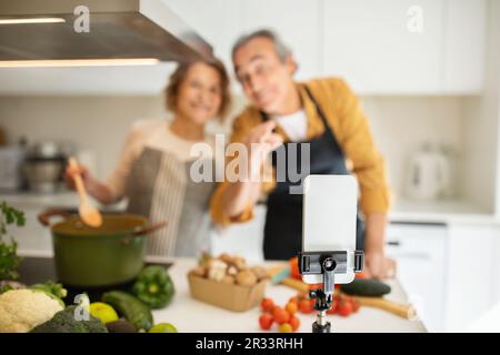 Selective focus on smartphone set on tripod over loving senior spouses cooking dinner together at modern kitchen Stock Photo