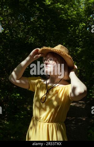 Beautiful Asian girl with glasses, straw hat and yellow dress in the forest in summer, beautiful shadows, sunlight, portrait. Dreamy young teen girl. Stock Photo