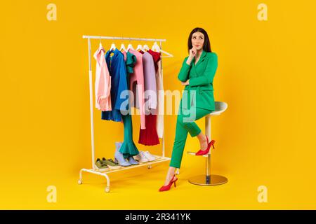 Pretty stylist lady sitting near garment rack and choosing new clothes, woman thinking about creating new outfits Stock Photo